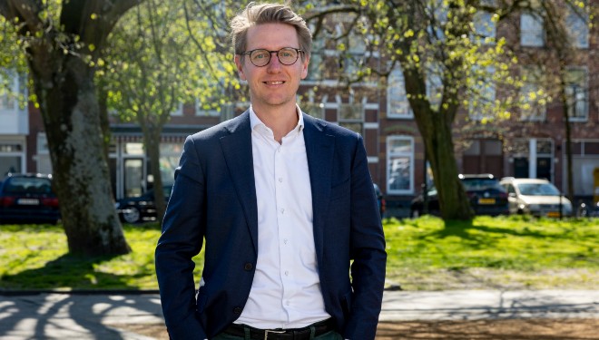 Portretfoto van Daniel van Drunen, buiten, op een zonnige dag. Daniel draagt een donkerblauw pak en wit overhemd zonder stropdas.